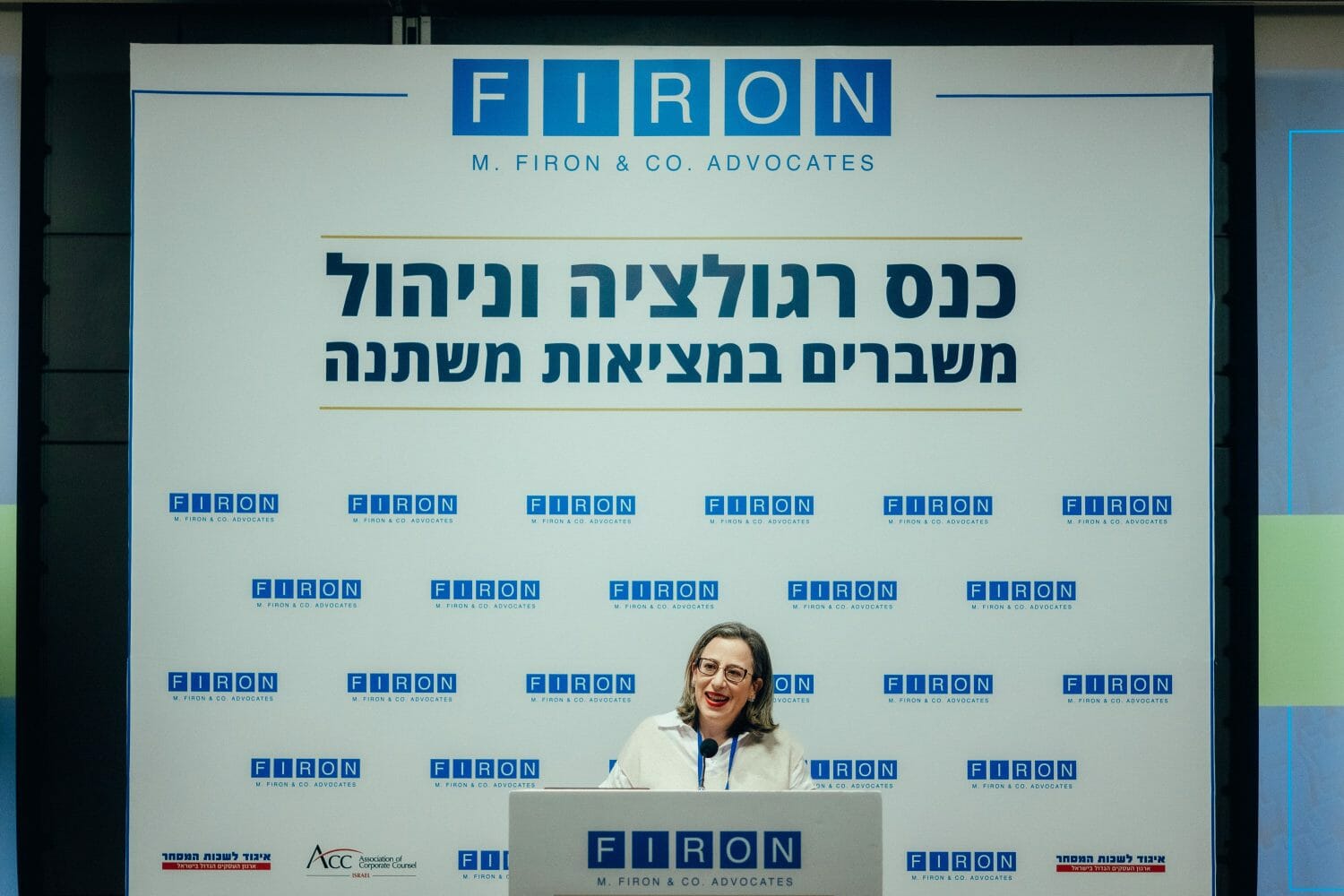 תמונה של אישה מדברת בפודיום נואמים כשמאחוריה רקע ממותג של כנס רגולציה וניהול משברים במציאות משתנה של פירון.